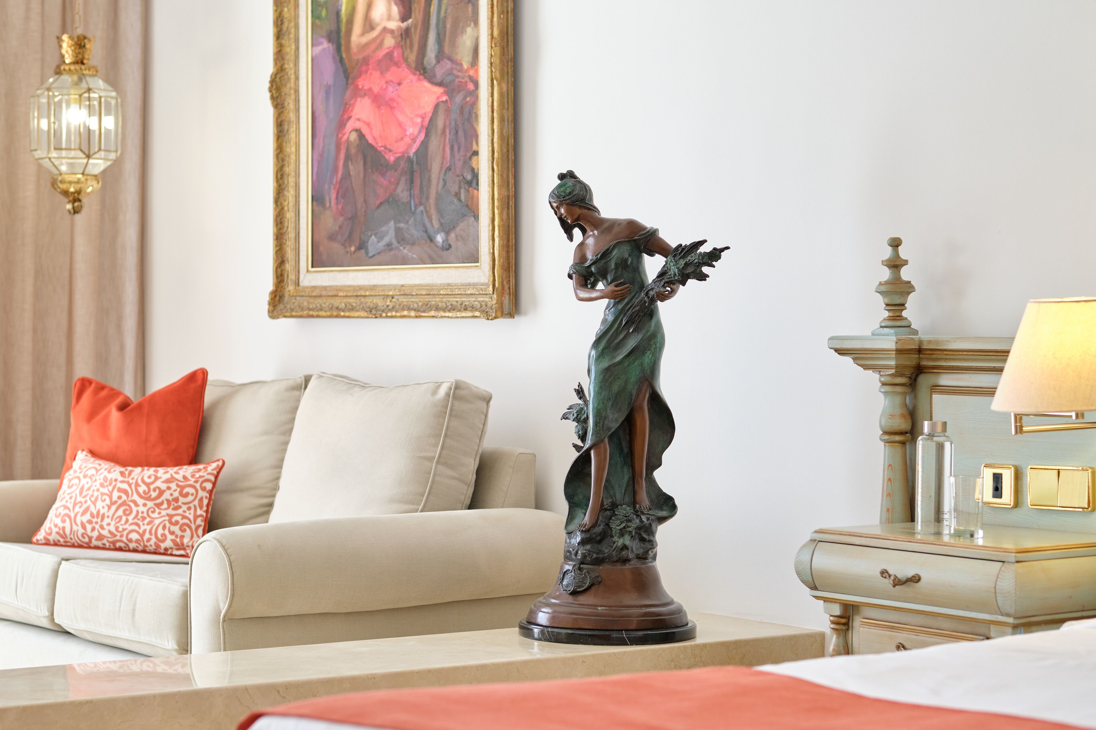 Ein Zimmer mit einer Statue auf einem Sockel, einer Couch mit gemusterten Kissen, einer Lampe und einem gerahmten Gemälde an der Wand.