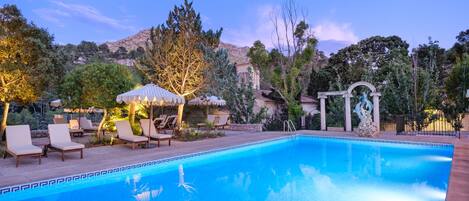 A swimming pool with lounge chairs and a gazebo, surrounded by trees and a mountainous backdrop.