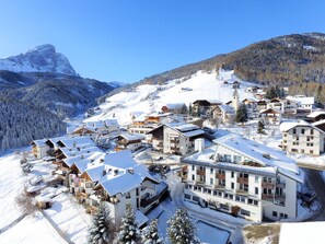 A snow-covered mountain resort with chalet-style buildings, ski slopes, and a prominent mountain peak in the background. - Hotel Pütia (San Martino in Badia)