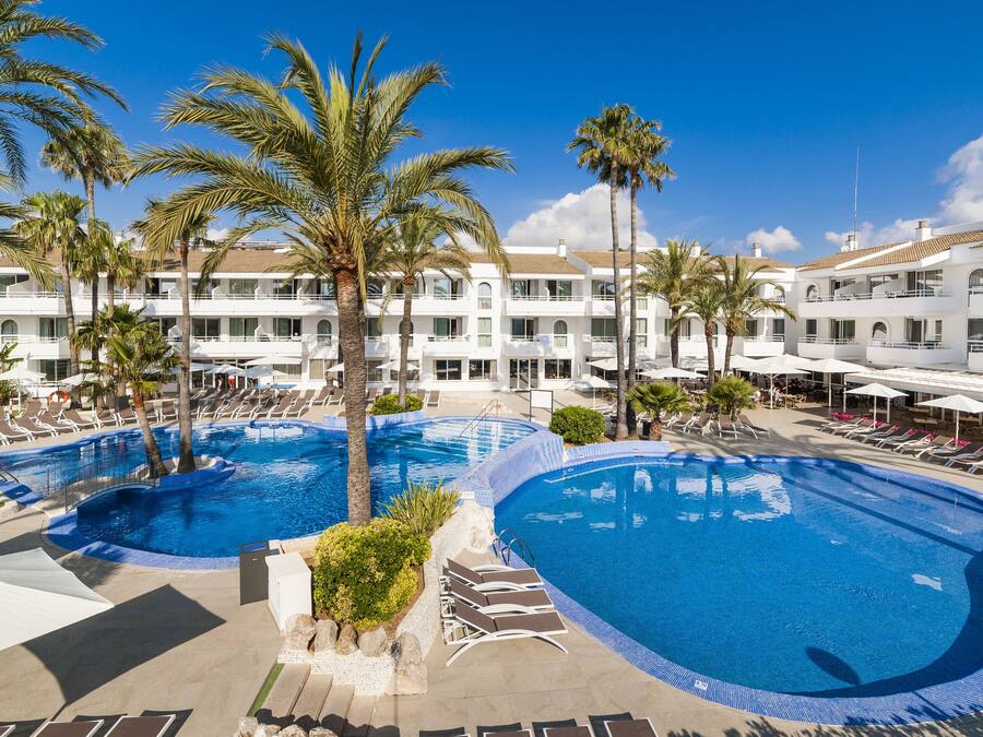 Um resort com duas piscinas espaçosas, palmeiras e um edifício branco com sacadas.