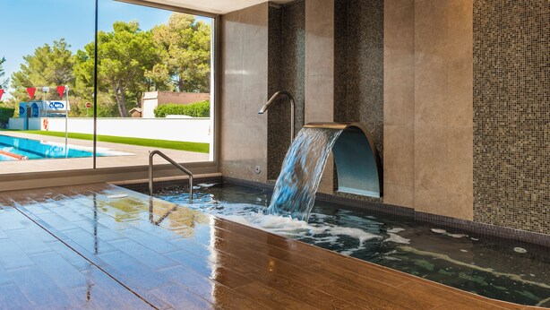 A modern indoor pool area with a waterfall feature, large windows, and a view of the outdoors. - Hoposa Apartamentos Montelin (Pollenca)