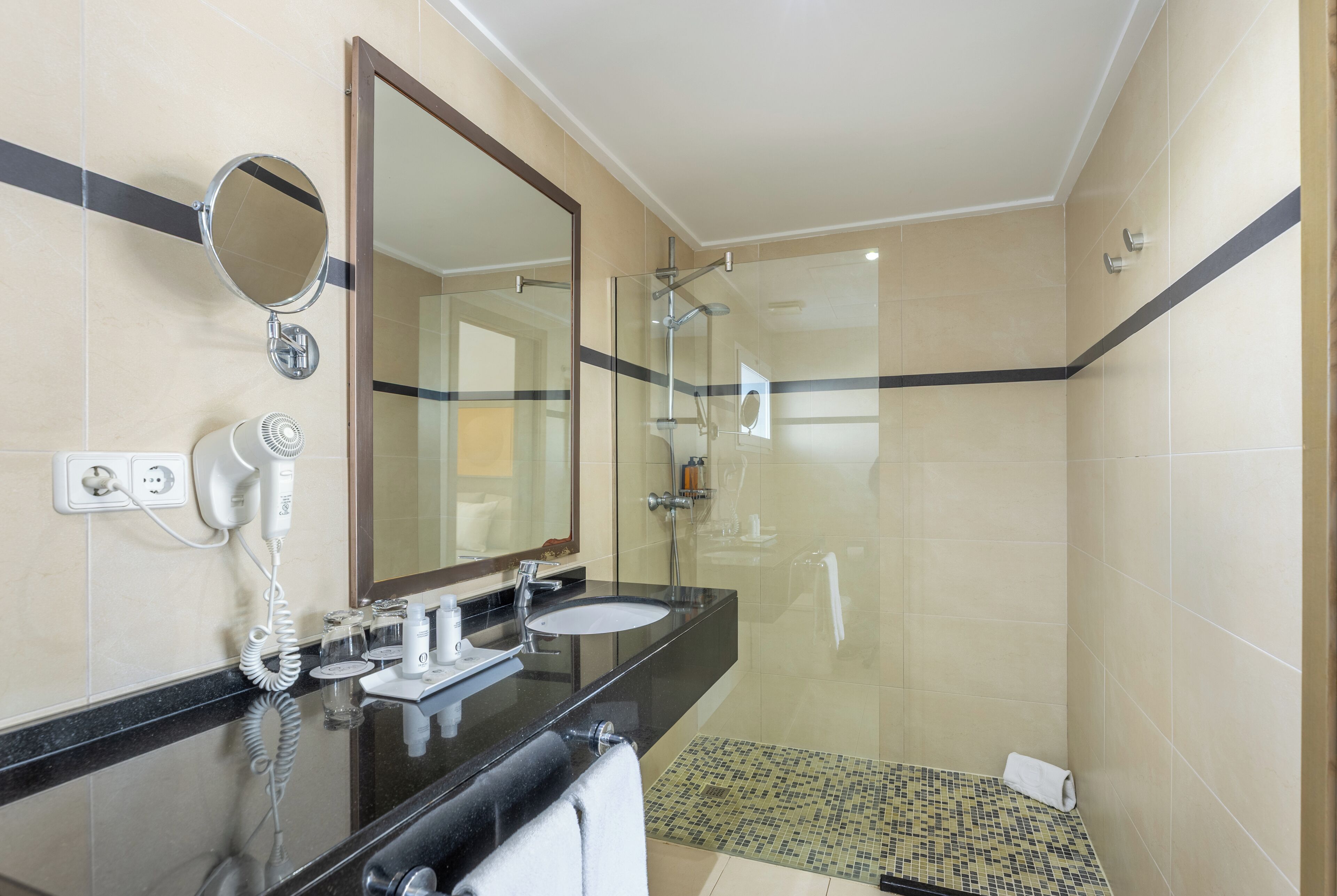 A modern bathroom with a black countertop, a round mirror, a glass-enclosed shower, and a tiled floor.