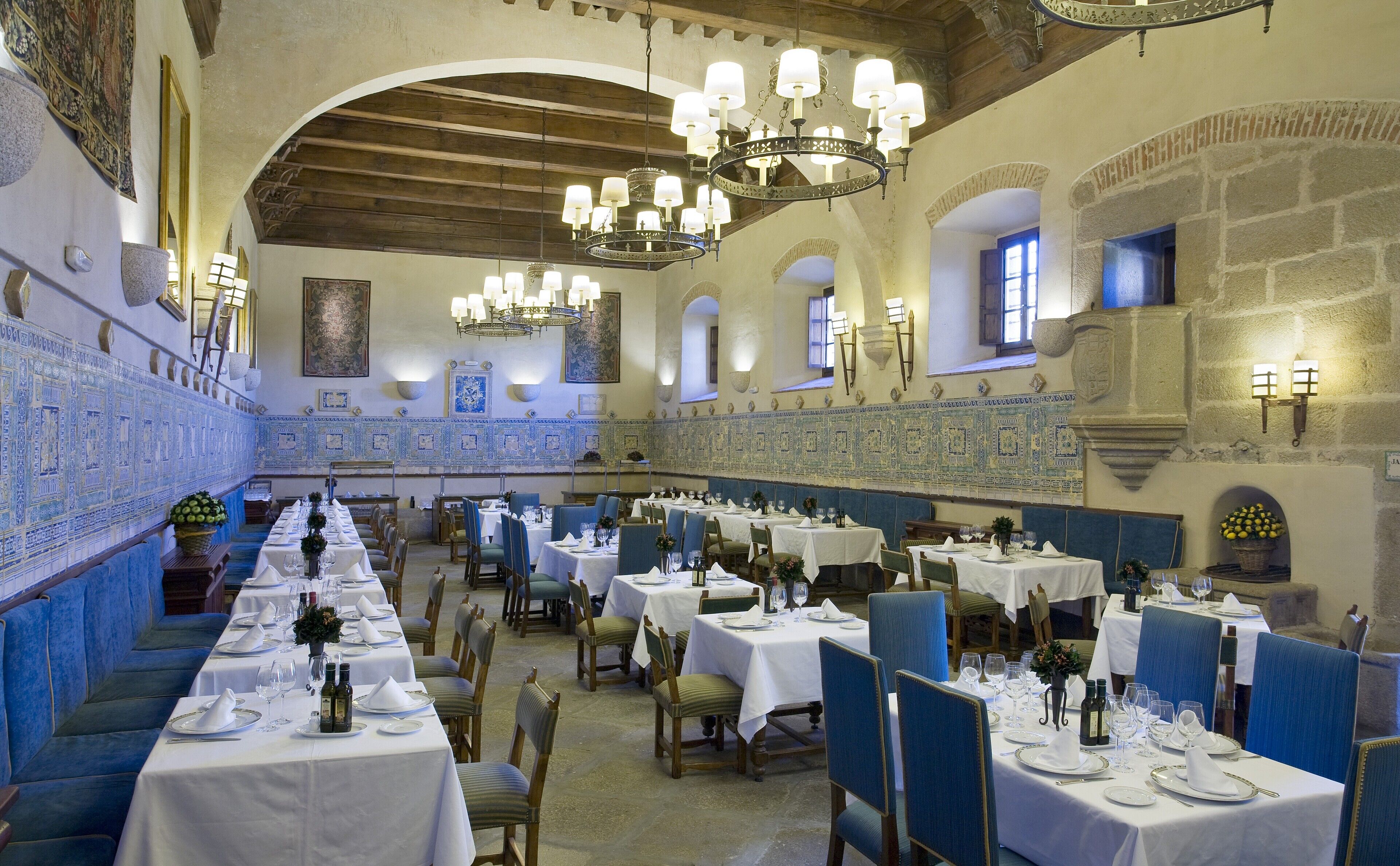 A dining area with multiple tables set for service, featuring a high ceiling with chandeliers and a stone wall with a fireplace.