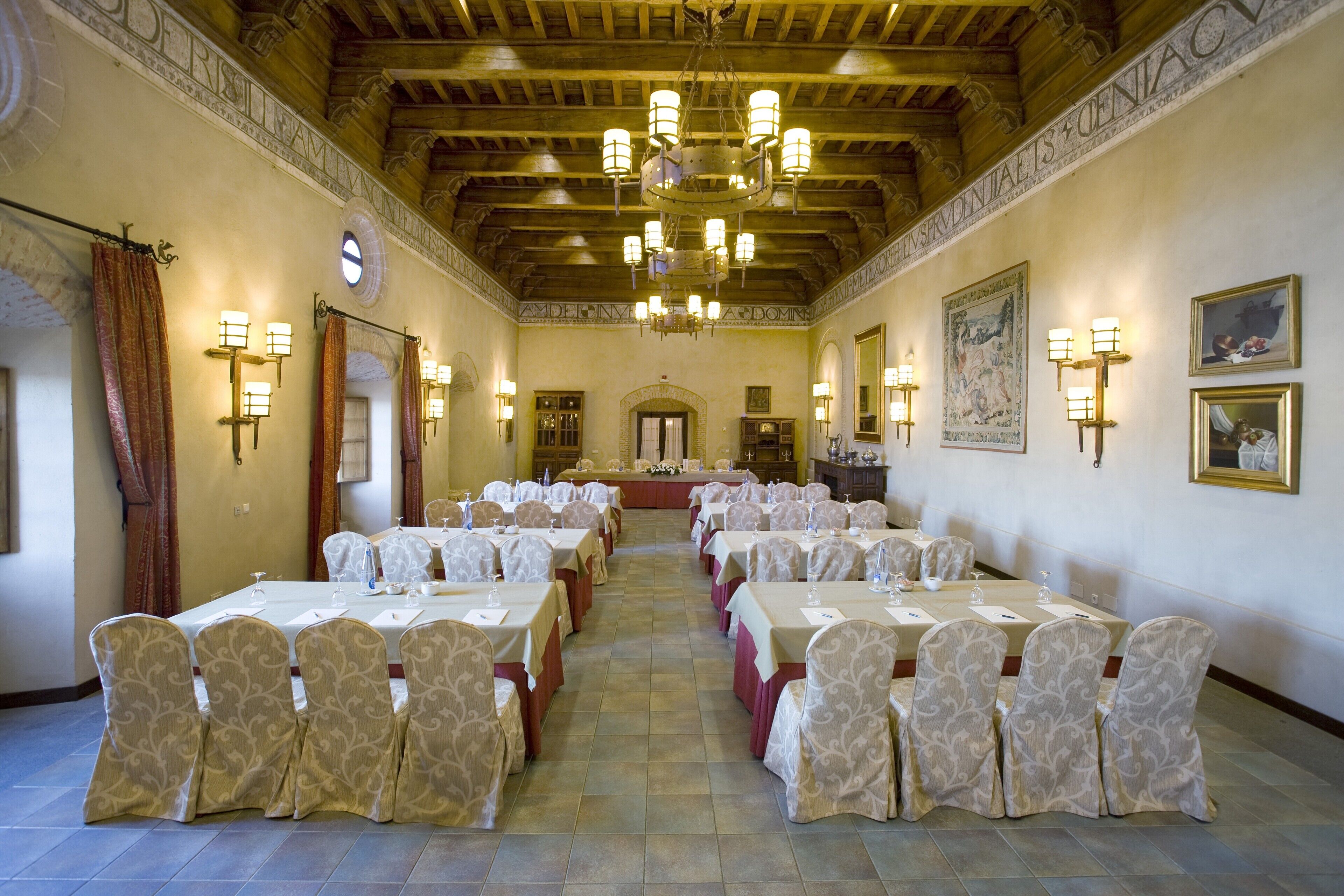 Een feestzaal met meerdere ronde tafels, elk gedekt met witte tafelkleden en beklede stoelen, en een kroonluchter aan het plafond.