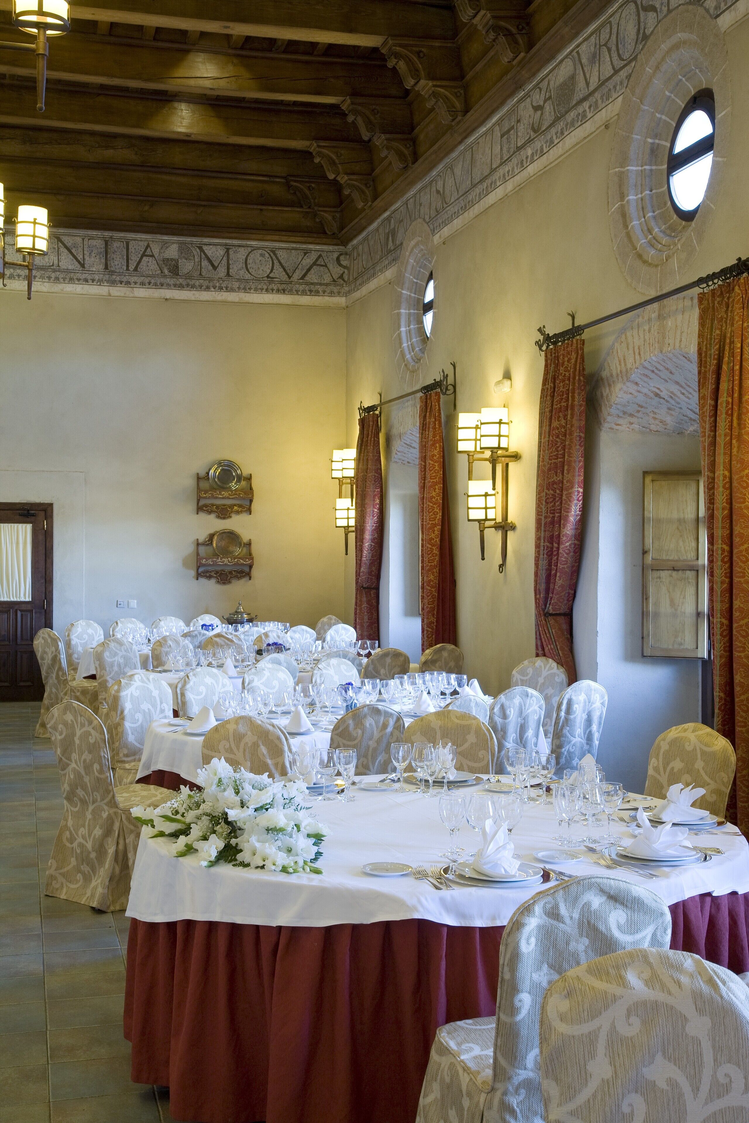 A banquet hall with round tables set for a formal event, featuring white tablecloths, floral centerpieces, and elegant chairs.