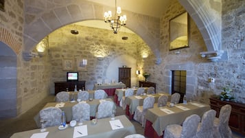 Un salón de conferencias con paredes de piedra, una araña, mesas con sillas y una televisión.