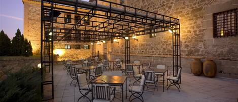 Un patio al aire libre con mesas y sillas, un edificio de piedra al fondo y una estructura de pérgola de metal.