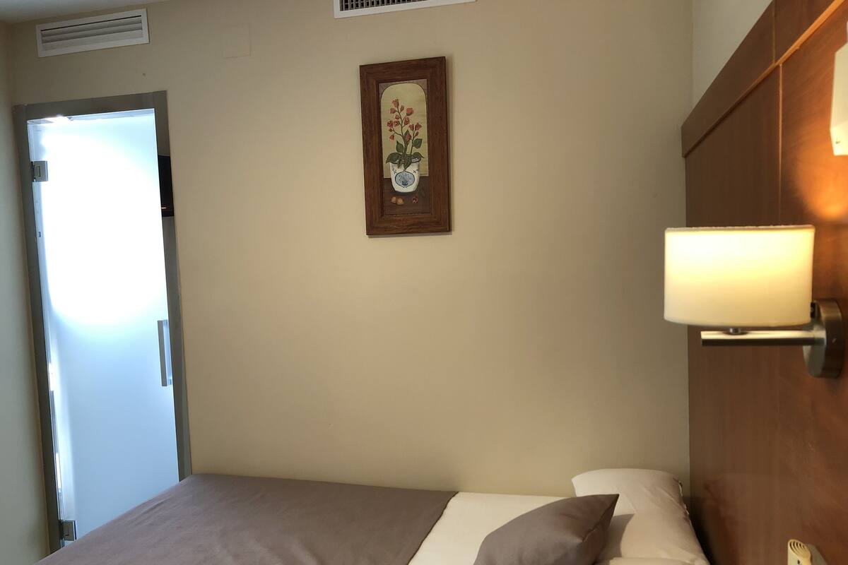 A hotel room with a bed, a nightstand, a wall-mounted lamp, a door, and a framed picture on the wall.