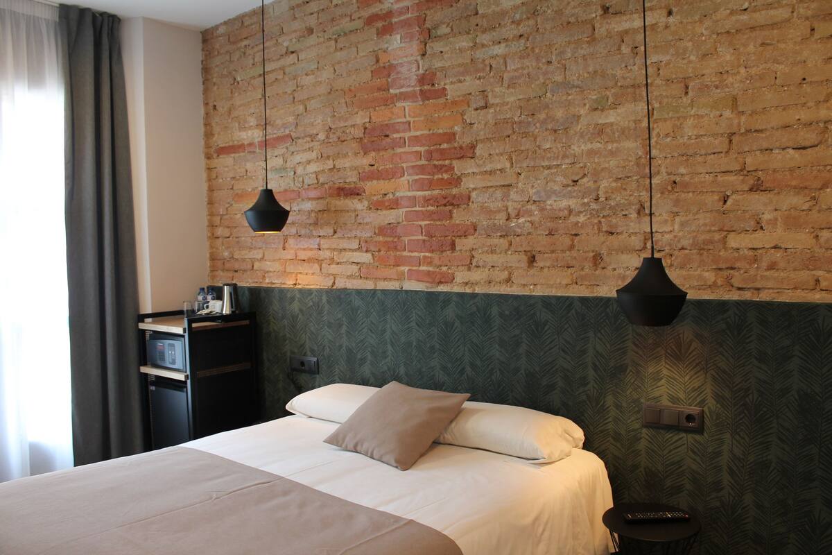 A hotel room with a brick wall, a bed with white linens, a small bedside table, and a hanging light fixture.
