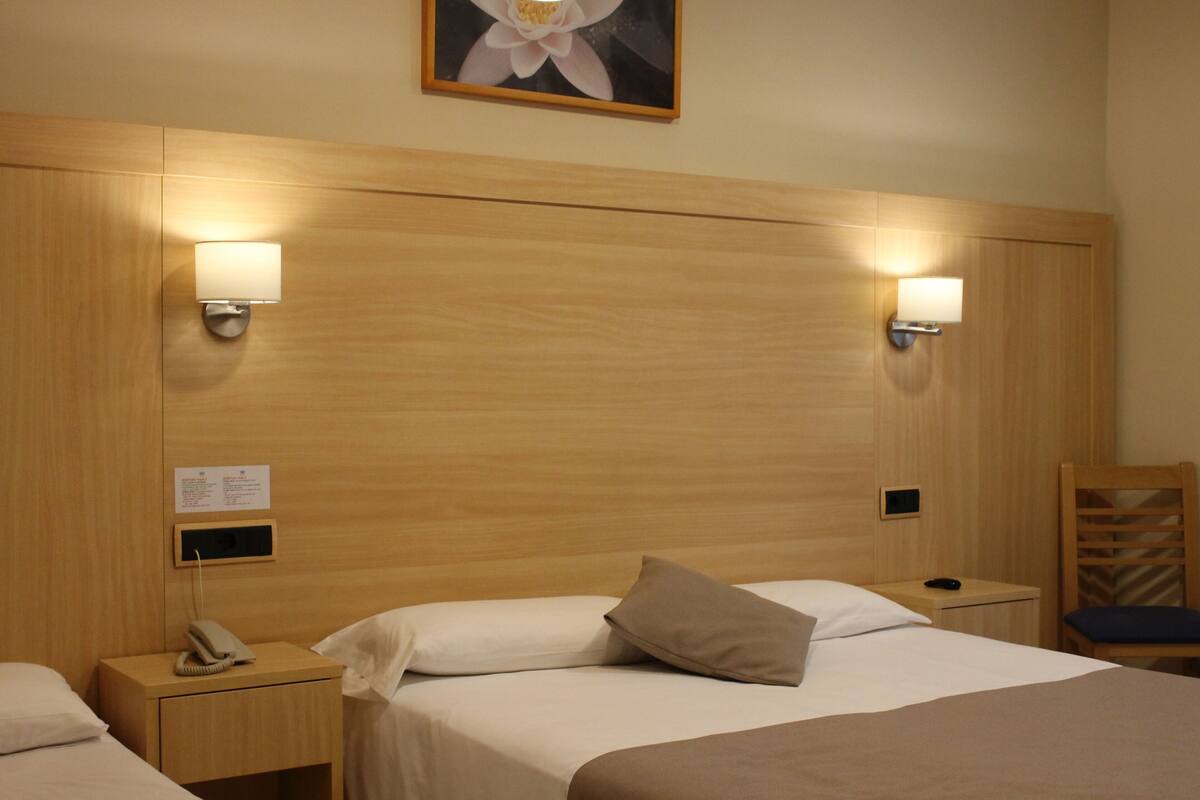 A hotel room with a wooden headboard, two bedside tables with lamps, a bed with white linens, a single pillow, a chair, and a framed picture on the wall.