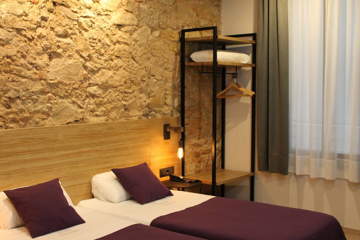 A hotel room with a stone wall, a wooden headboard, two beds with purple bedding, a hanging wardrobe, and a bedside table with a lamp.