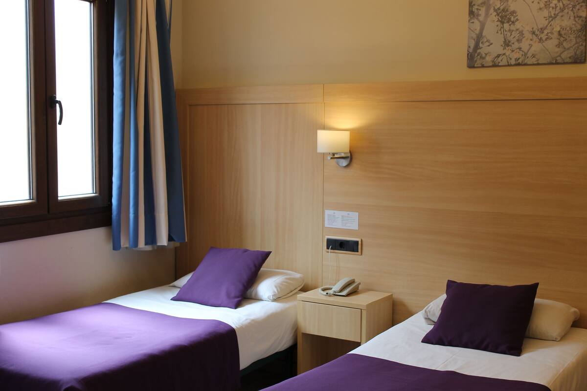A hotel room with two beds, each with a purple bedspread and pillow, a bedside table with a lamp, and a window with curtains.