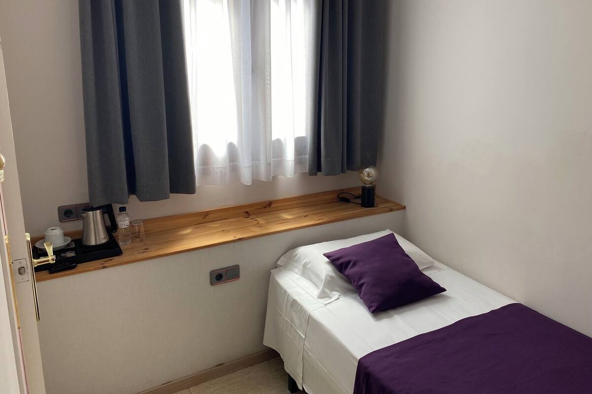 A small, neatly arranged bedroom with a single bed, a nightstand, a window with curtains, and a wall-mounted light.