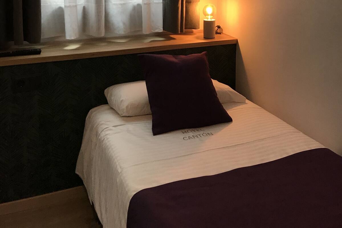 A hotel room with a bed, a nightstand with a lamp, and a window with curtains.