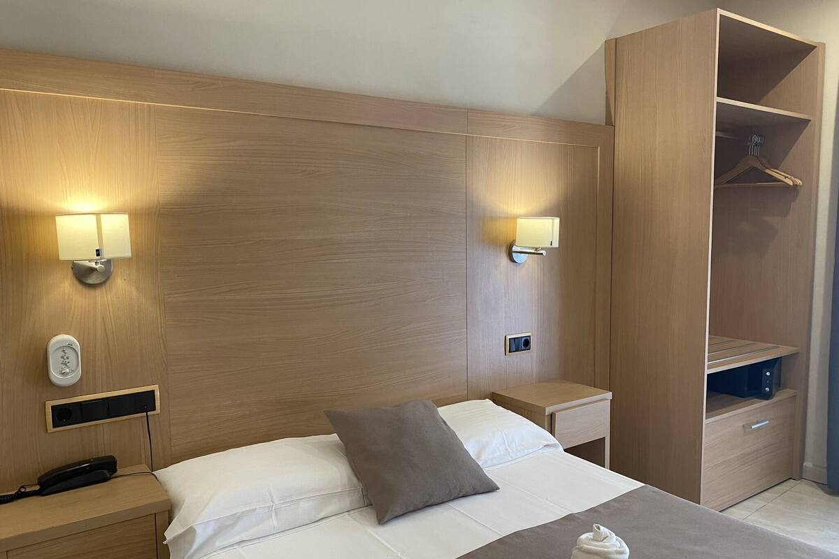 A hotel room with a wooden headboard, bedside lamps, a telephone, a bedside table, a bed with white and grey bedding, and a wardrobe with a hanging space.