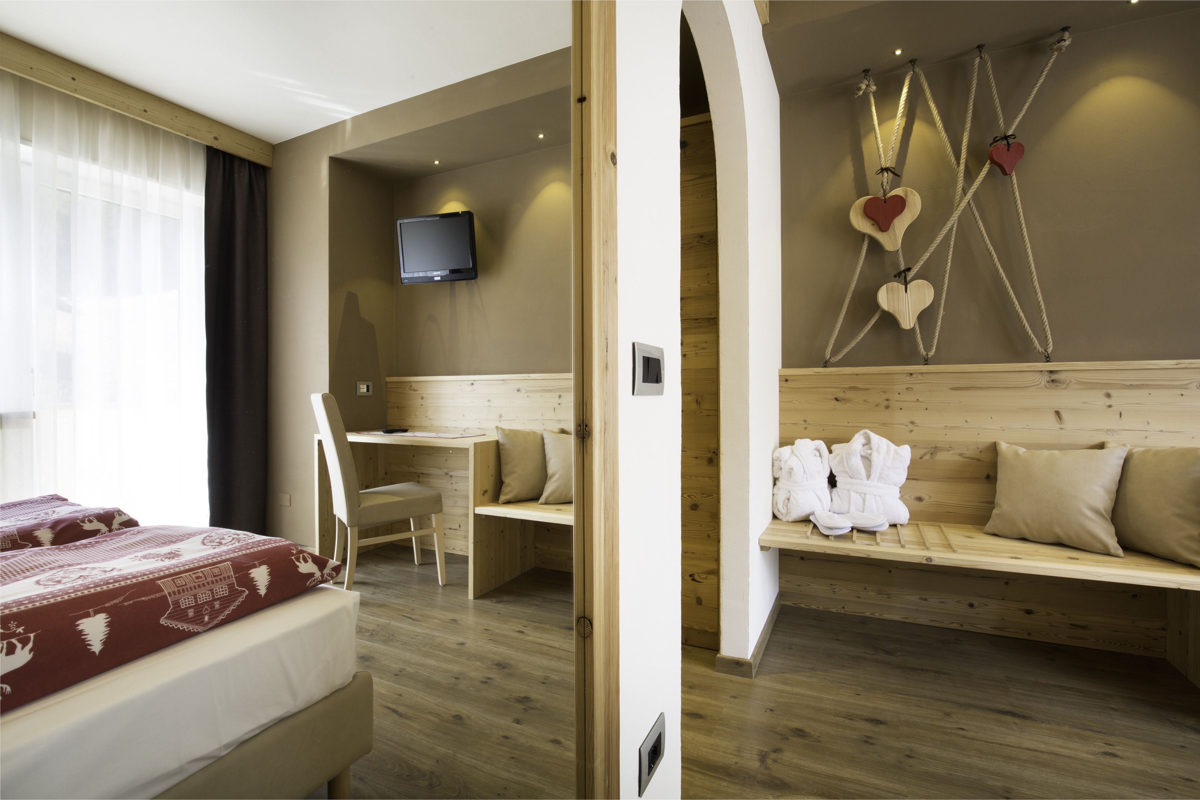 Quarto de hotel com cama, escrivaninha, televisão, um banco com toalhas e uma parede decorada com corações.