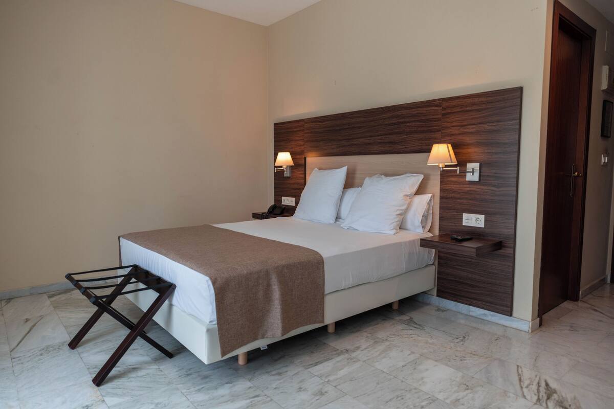 Una habitación de hotel moderna con una cama grande, cabecera de madera, lámparas de noche y suelo de mármol.