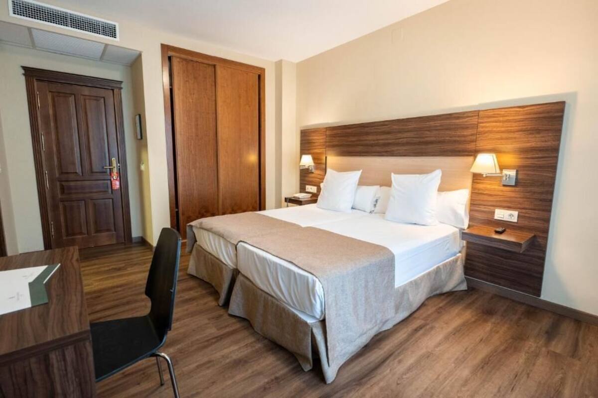 Habitación de hotel con cama doble, dos mesitas de noche con lámparas, armario de madera, escritorio con silla y una pequeña mesa con lámpara.