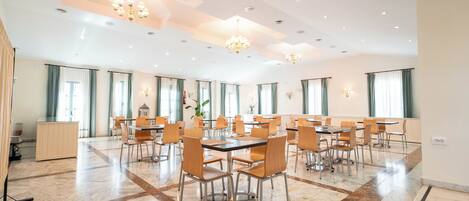 A spacious dining area with multiple tables and chairs, large windows with curtains, and a chandelier.