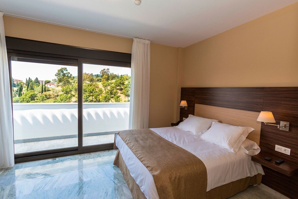 Une chambre d’hôtel avec un lit, une table de chevet, une lampe et une vue sur la verdure grâce à une grande fenêtre.