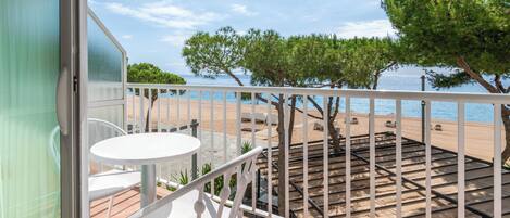 Un balcon avec une table et des chaises blanches, donnant sur une plage bordée d’arbres et sous un ciel dégagé.