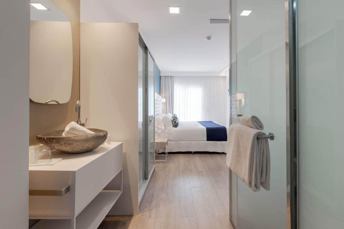 A modern hotel room with a large round sink, a bed with a blue accent, and a glass shower enclosure.