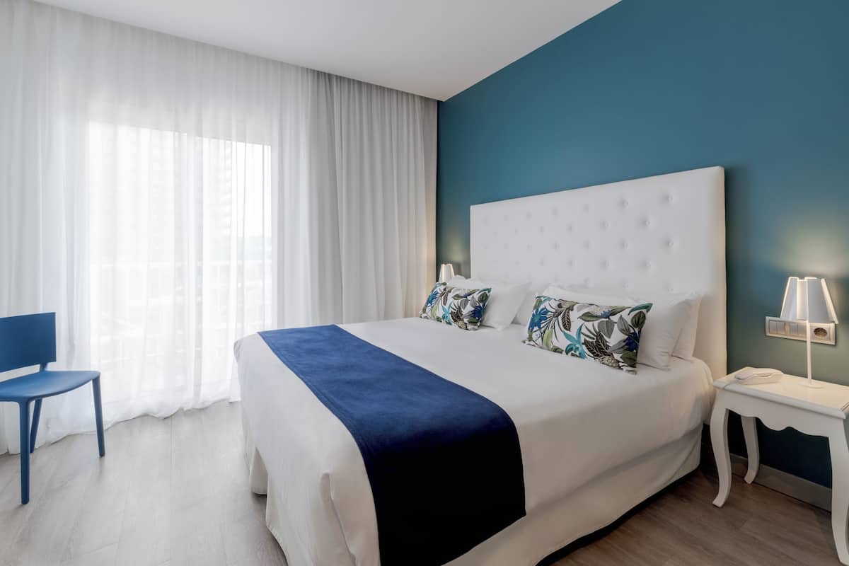 A modern hotel room with a large bed, a blue chair, a bedside table with a lamp, and a view of the outdoors through sheer curtains.