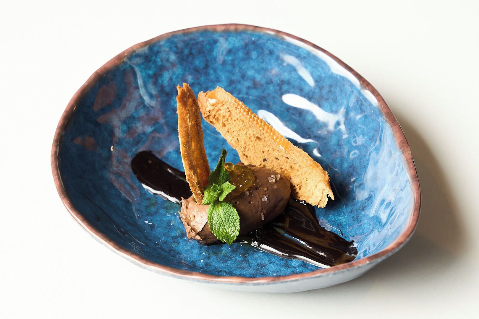 A blue ceramic bowl containing a dessert with a chocolate element, a piece of bread, and a mint leaf.