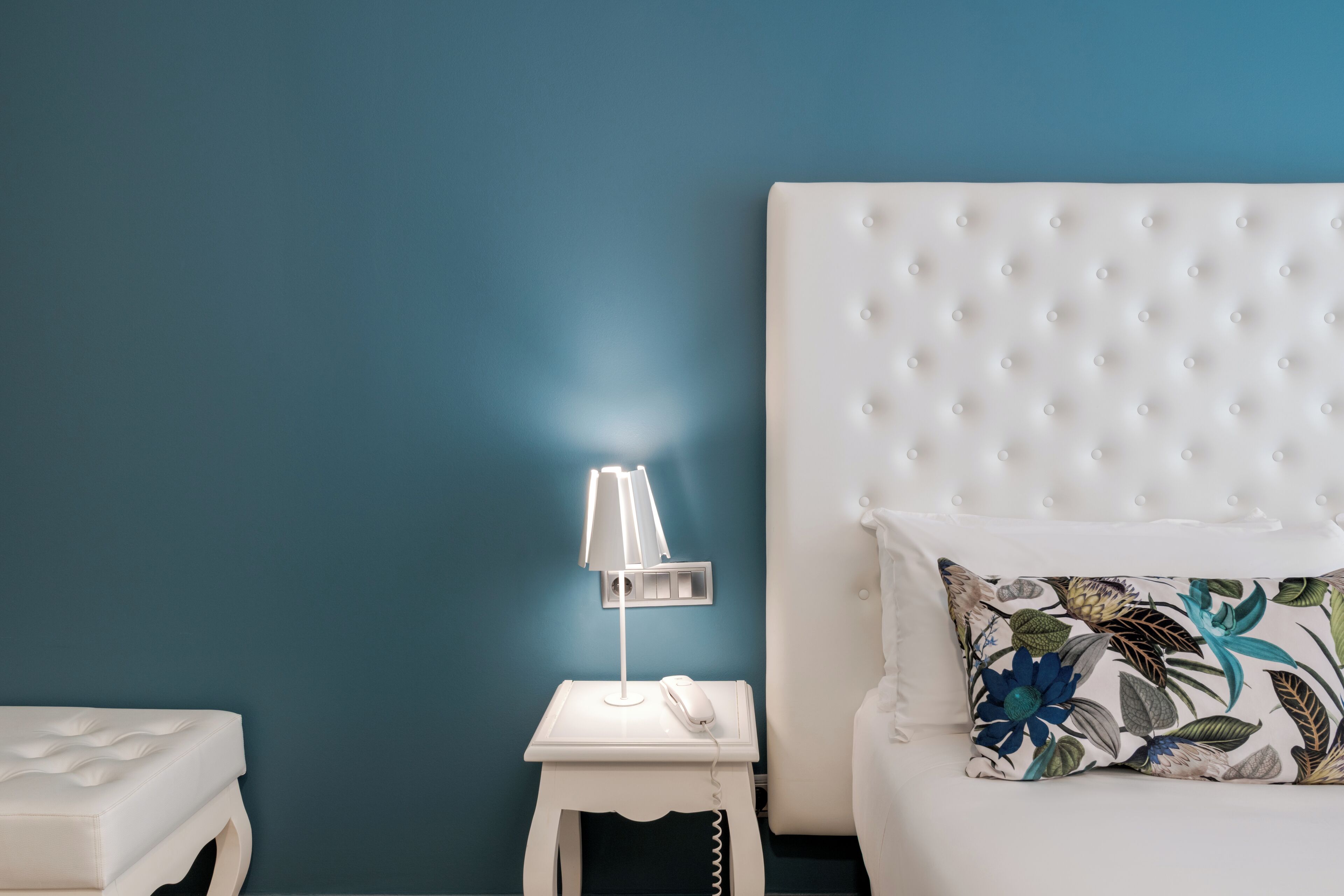 Een slaapkamer met een gestuukt hoofdeinde, een witte nachtkastje met een lamp, een bloemenkussen en een witte matras.