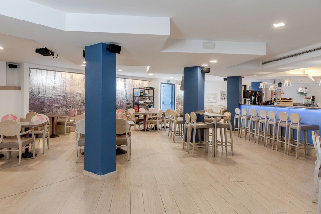 A spacious dining area with multiple tables and chairs, a bar area with stools, and a large window with a view of trees.