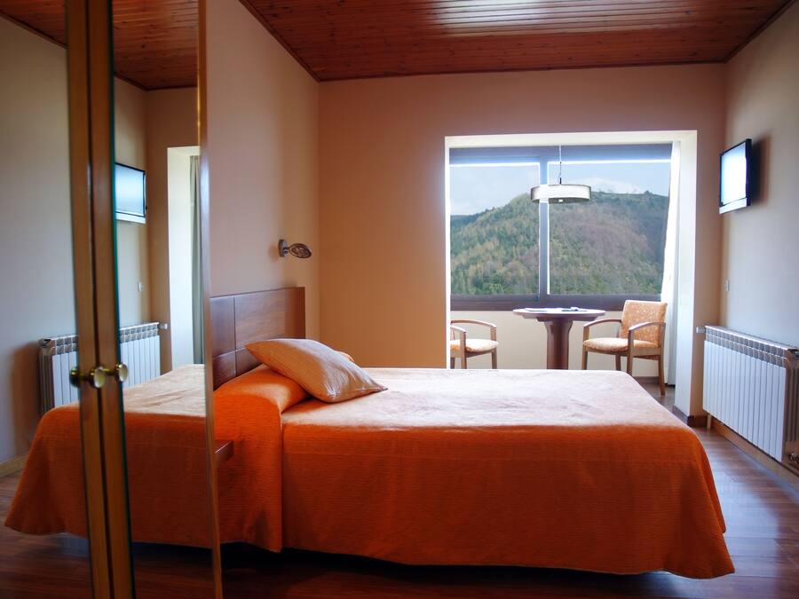 A bedroom with a bed, a window with a view of a mountain, a table, and two chairs.