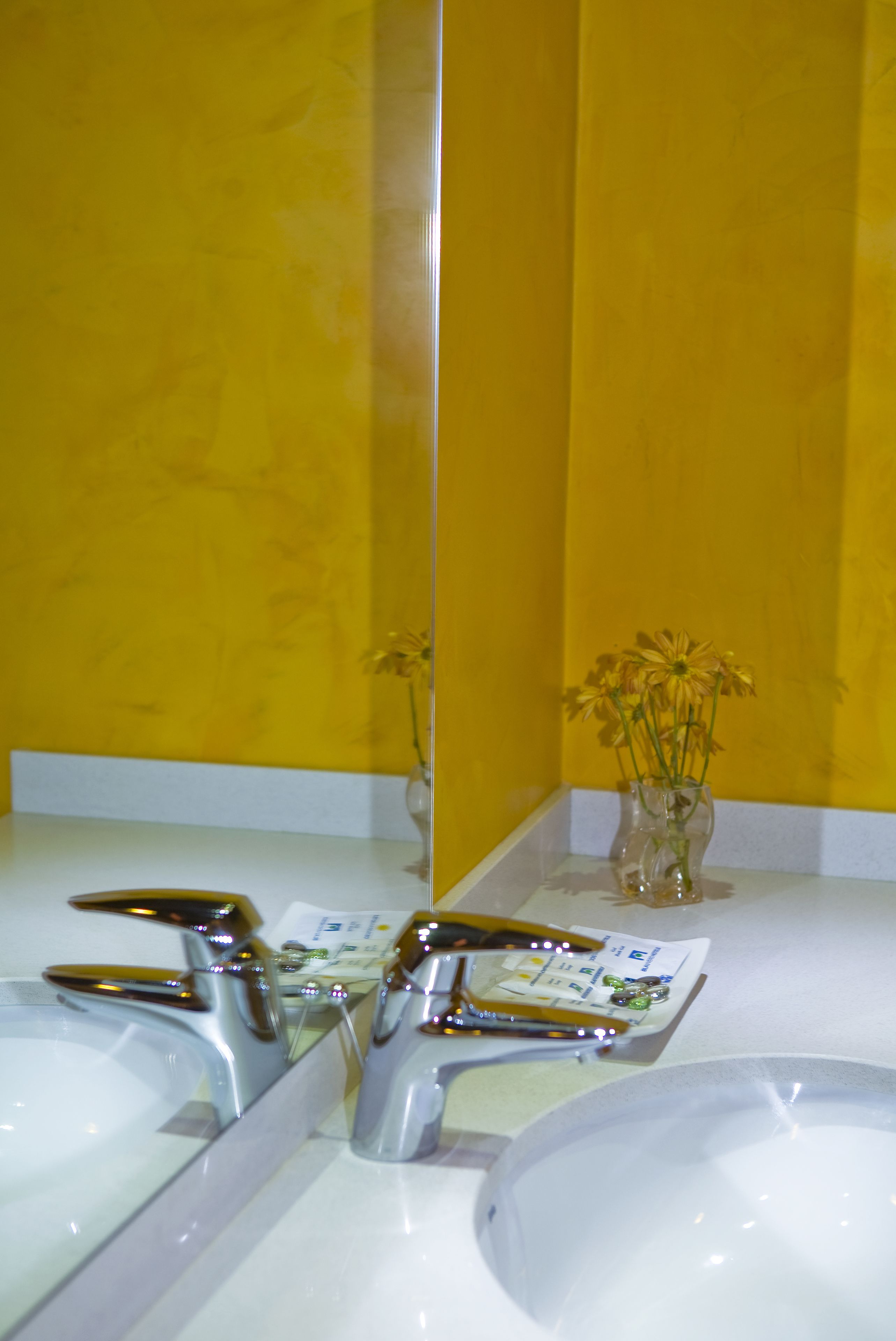 A bathroom with a yellow wall, a white sink, and a mirror reflecting the sink and a vase with flowers.