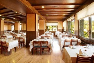 Restaurante com várias mesas preparadas para refeições, vigas de madeira no teto e grandes janelas que oferecem vista para o exterior.