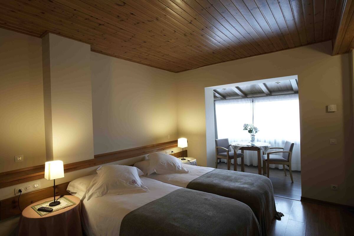 A hotel room with two beds, wooden ceiling, a small table with chairs, and a window with natural light.