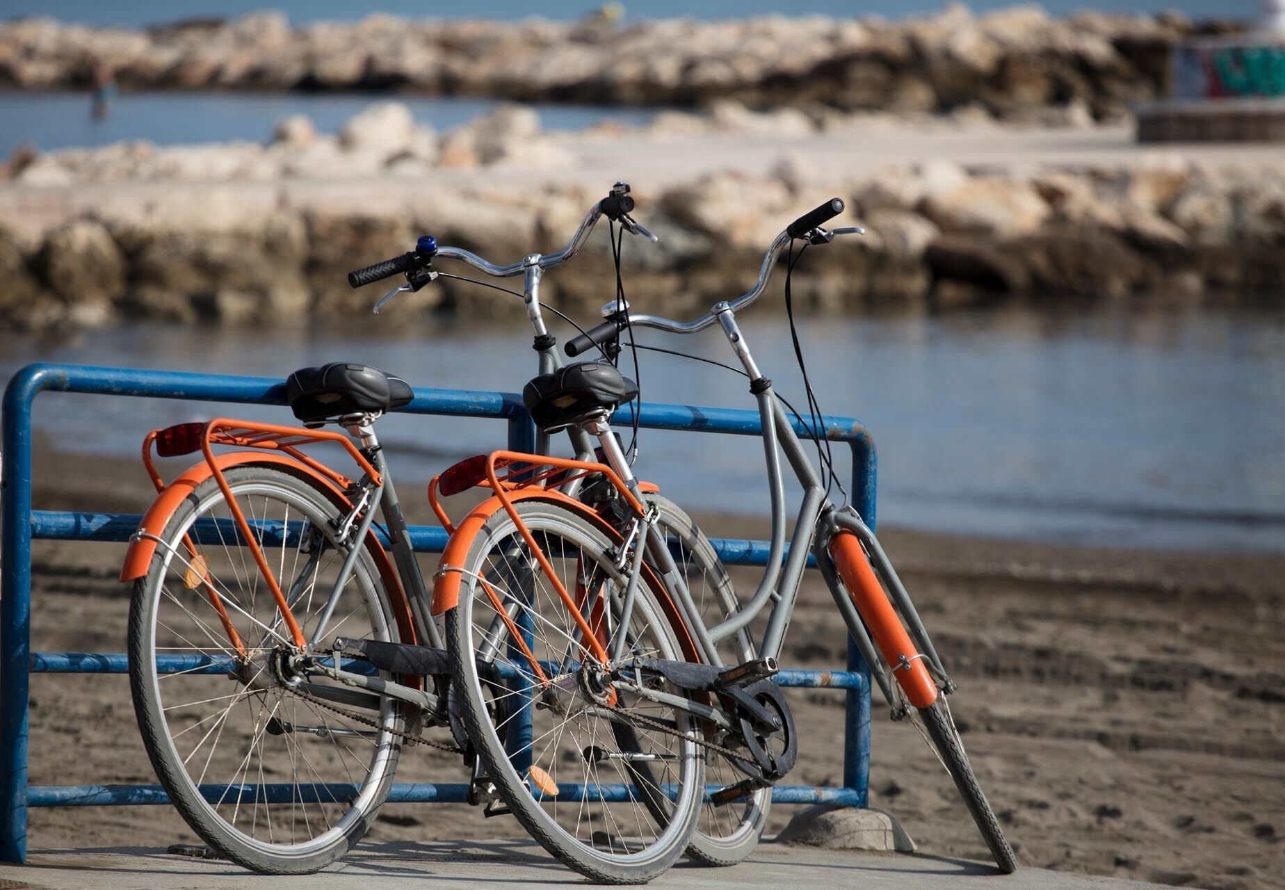 Dos bicicletas estacionadas frente a una barrera metálica azul, junto a una playa de arena con costa rocosa al fondo.