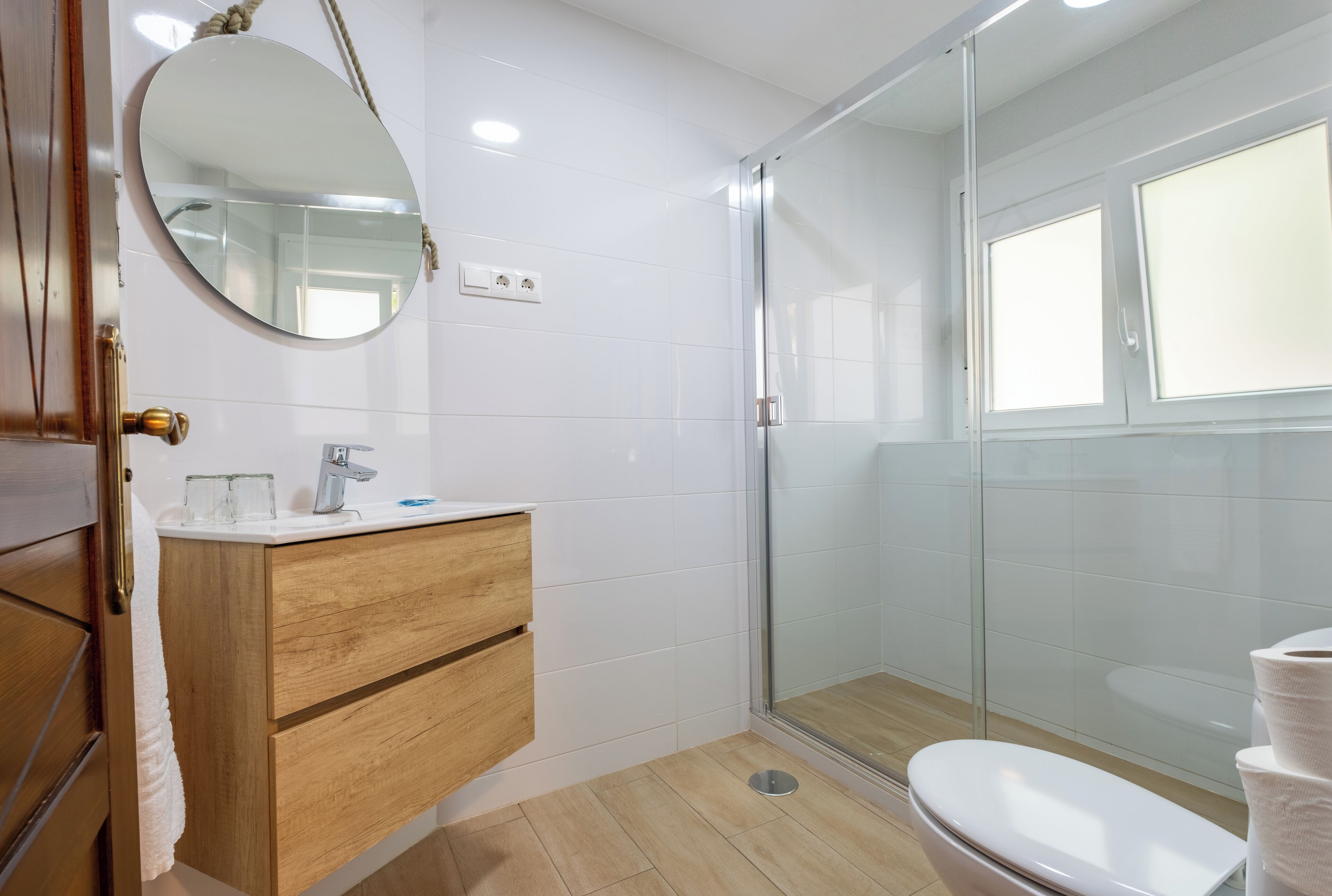 A modern bathroom with a wooden vanity, a round mirror, a glass-enclosed shower, and a toilet.