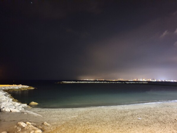 A nighttime beach scene with calm waters, a rocky shoreline, and distant city lights.
