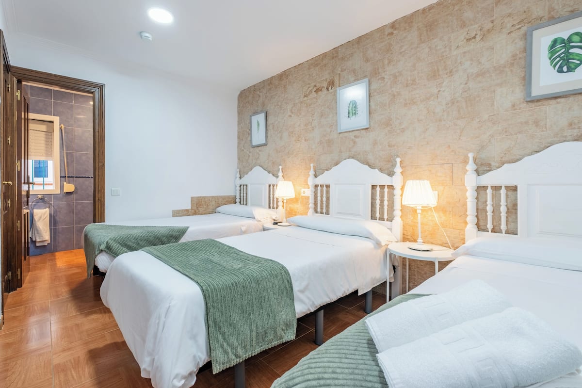A hotel room with three beds, each with white headboards and green throw blankets. There are framed pictures on the wall and a small table with a lamp between the beds.