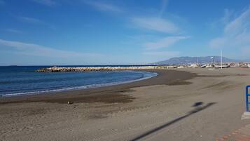 Una playa con un espigón rocoso, mar tranquilo y montañas a lo lejos.