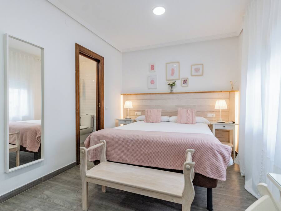 Un dormitorio con cama, mesitas de noche, un banco, un espejo y cuadros en la pared.