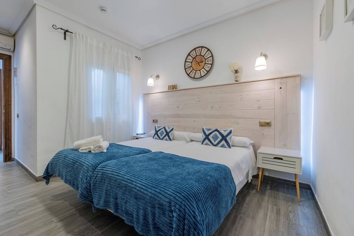 A double bed with blue bedding, a wooden headboard, a wall clock, and a bedside table with a lamp.