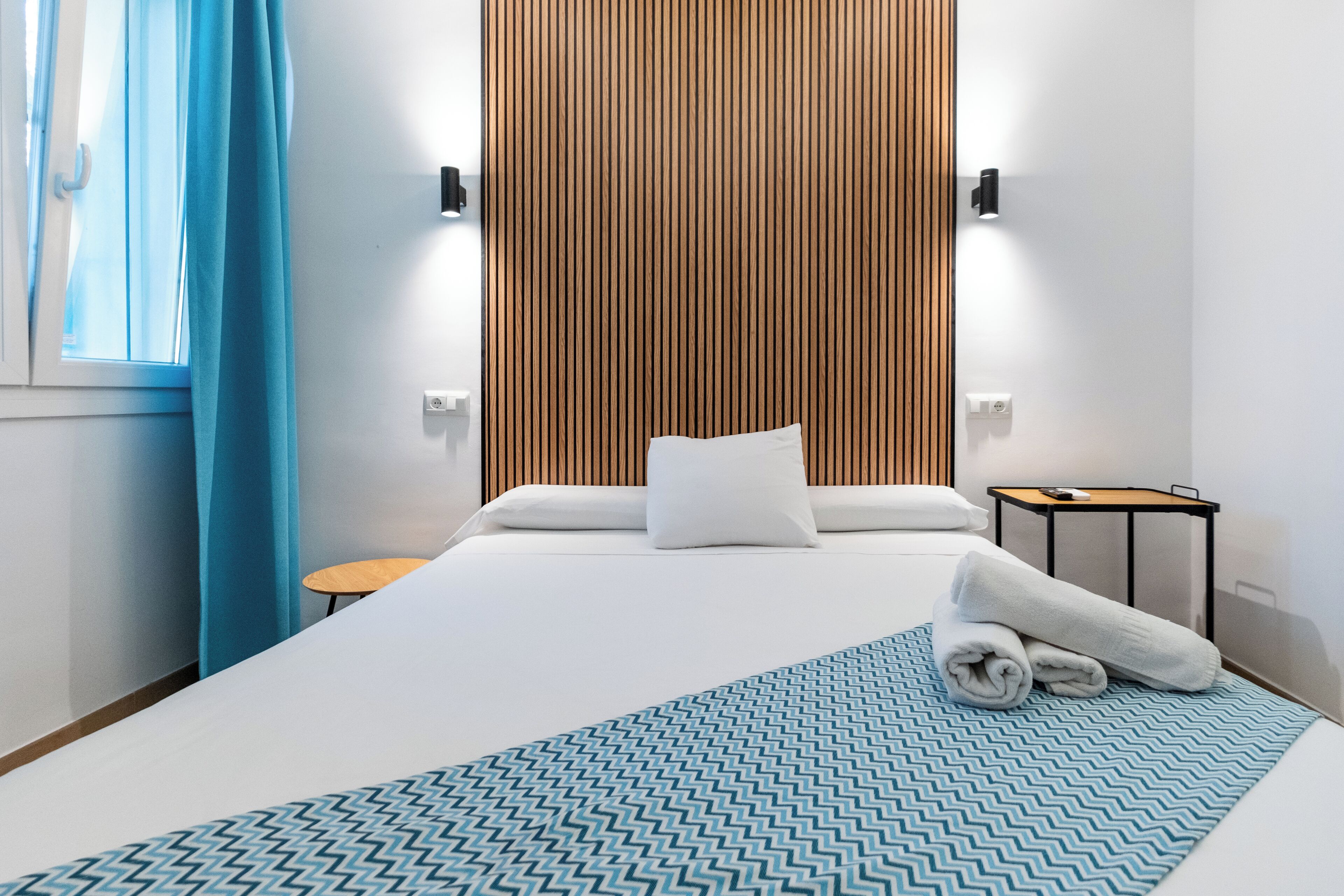 A hotel room with a large bed, a wooden panel wall, a small round table, and a blue patterned throw.