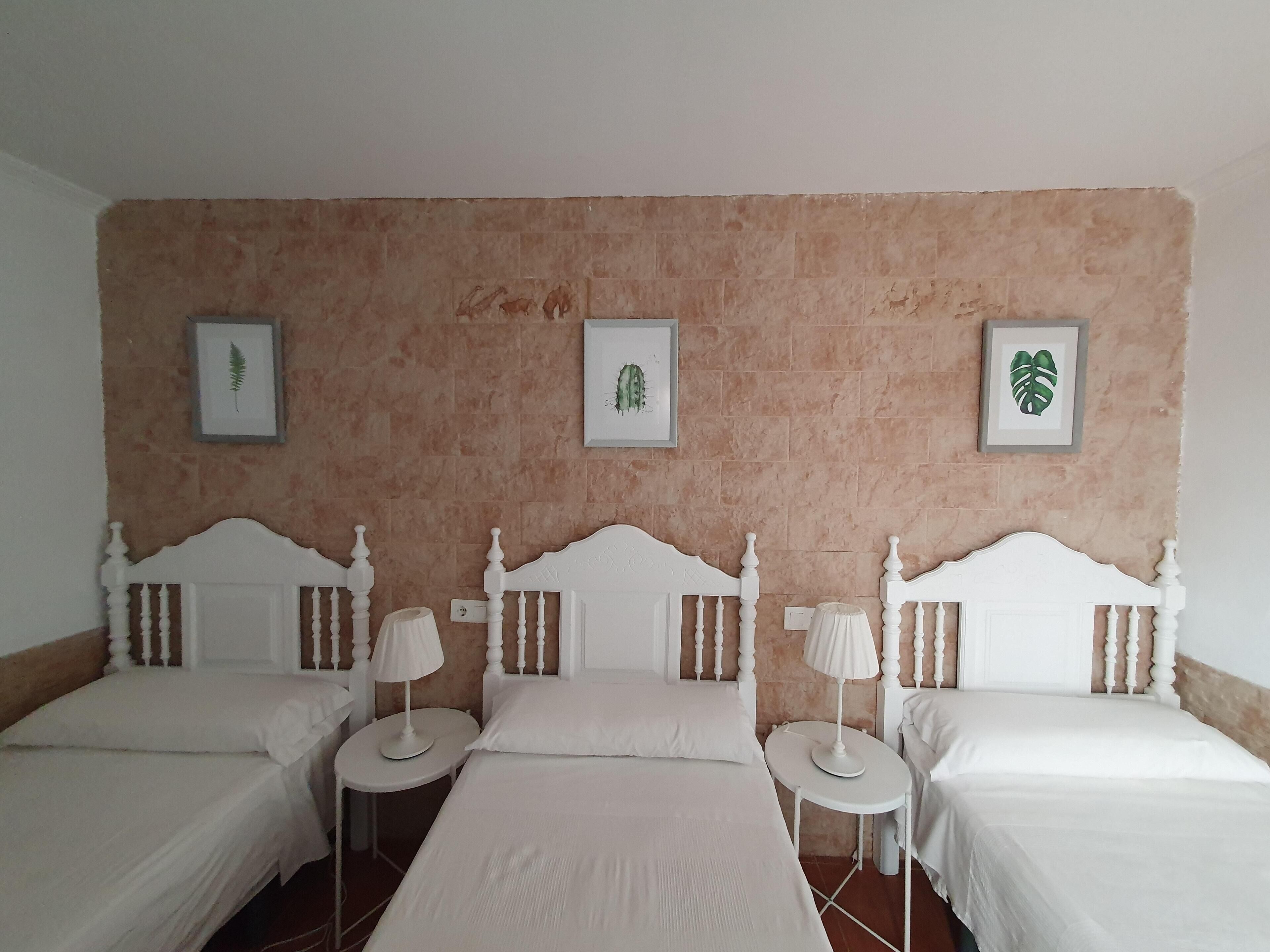 Habitación de hotel con tres camas individuales, cada una con cabecero blanco y mesitas de noche a juego. Las paredes son color beige y hay impresiones botánicas enmarcadas en la pared.