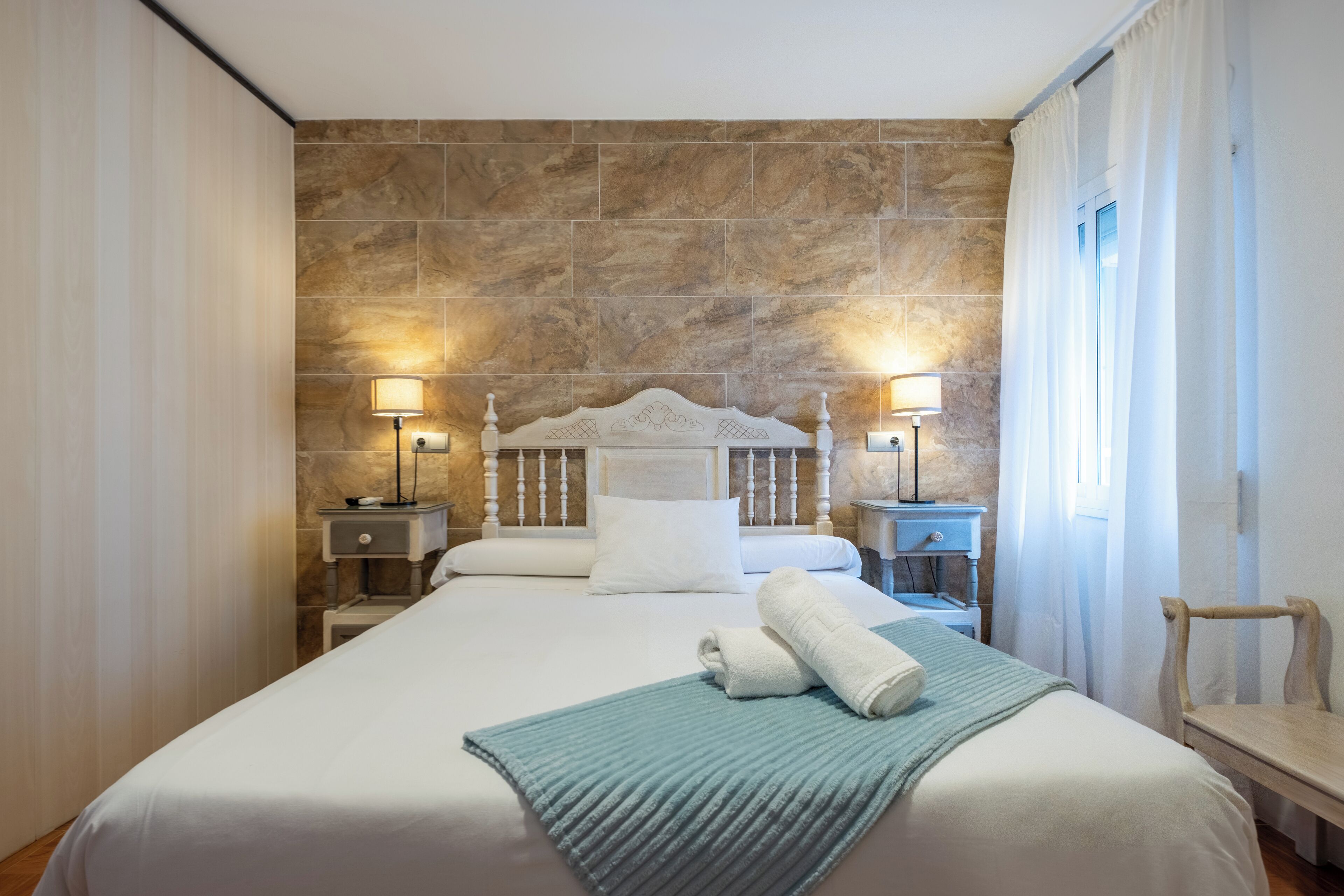 A hotel room with a stone wall, a white bed with a wooden headboard, two bedside tables with lamps, and a window with curtains.
