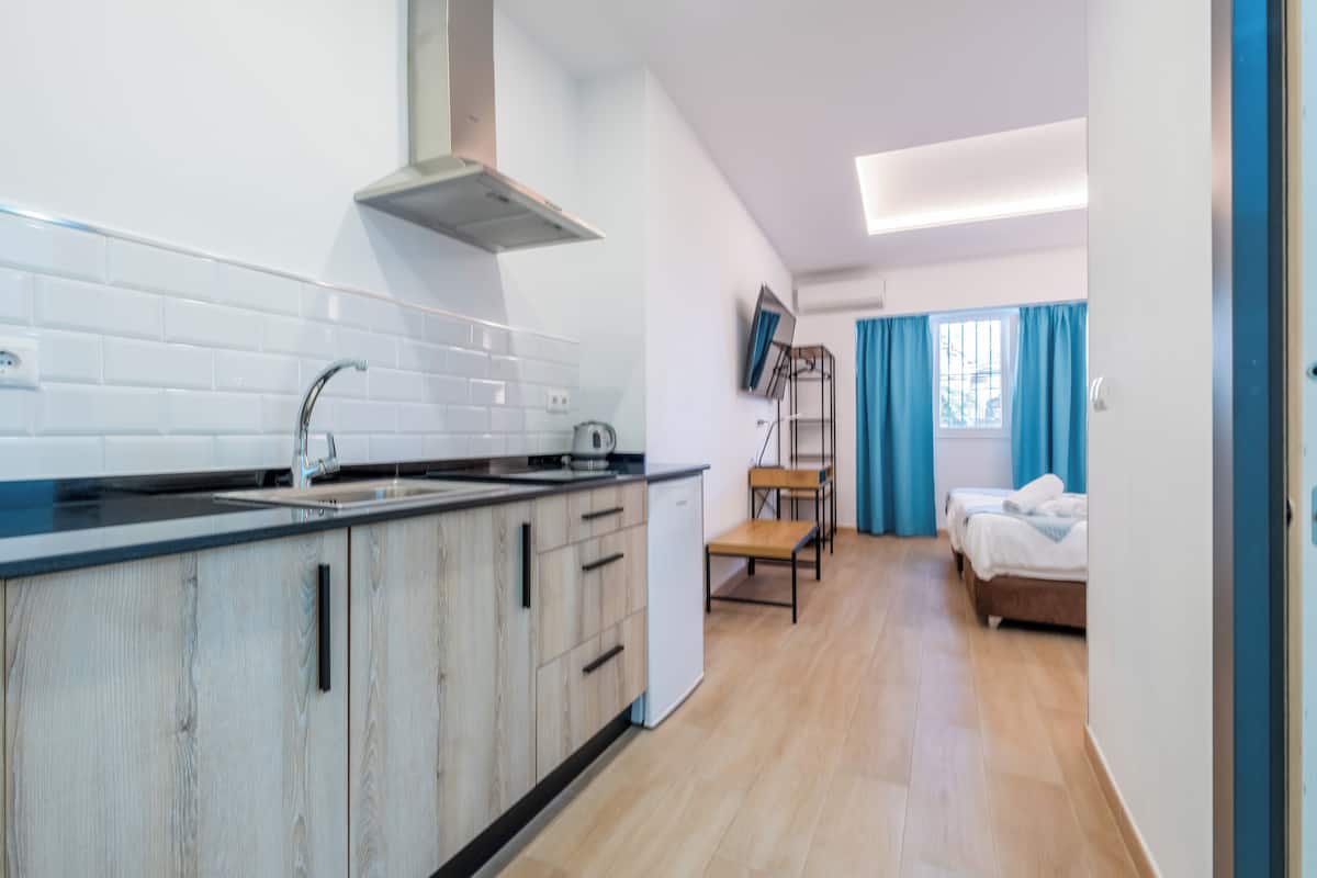 A modern kitchen with white tiles, a stainless steel range hood, and wooden cabinets. There is a dining area with a table and chairs, and a bedroom with a bed and curtains.