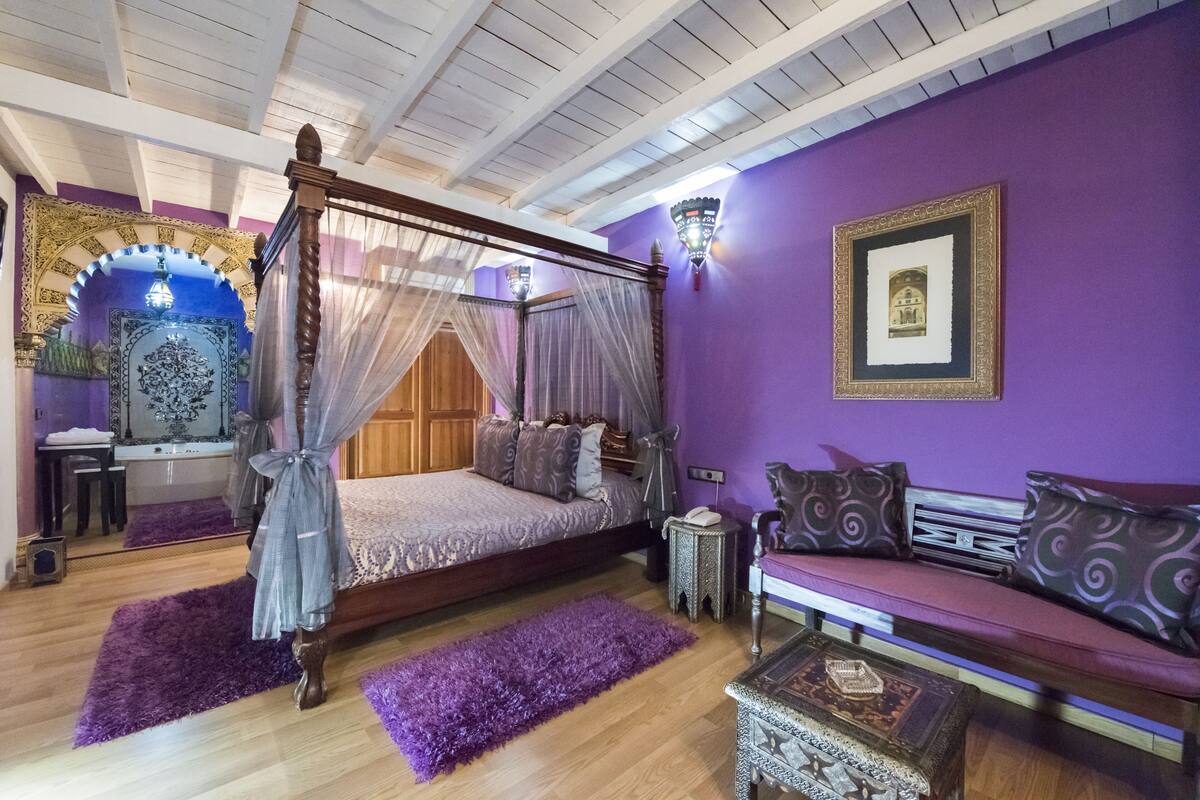 Ein Schlafzimmer mit Himmelbett, einem violetten Sofa und einem dekorativen Spiegel.