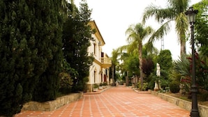 Un chemin pavé mène à un bâtiment doté d’un balcon, qui est entouré d’une végétation luxuriante et d’un mur de pierre.