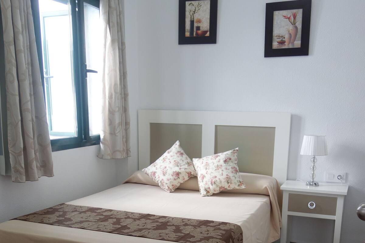 A neatly made bed with floral pillows, a bedside table with a lamp, and two framed pictures on the wall.