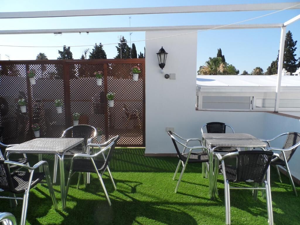 An outdoor patio with a table and chairs, a wooden fence with potted plants, and a white wall with a wall-mounted lamp.