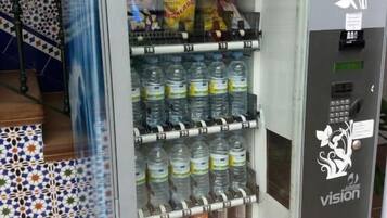 Máquina expendedora con diversas bebidas y aperitivos, ubicada en un área con baldosas.